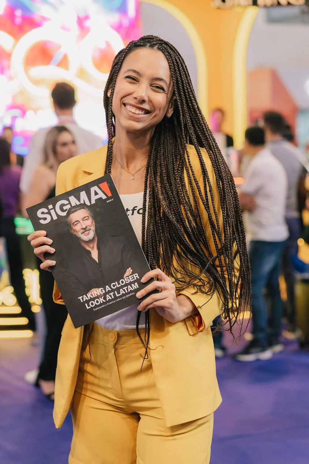 Mulher sorridente de cabelos trançados longos segurando uma revista com homem na capa, em ambiente com várias pessoas ao fundo.