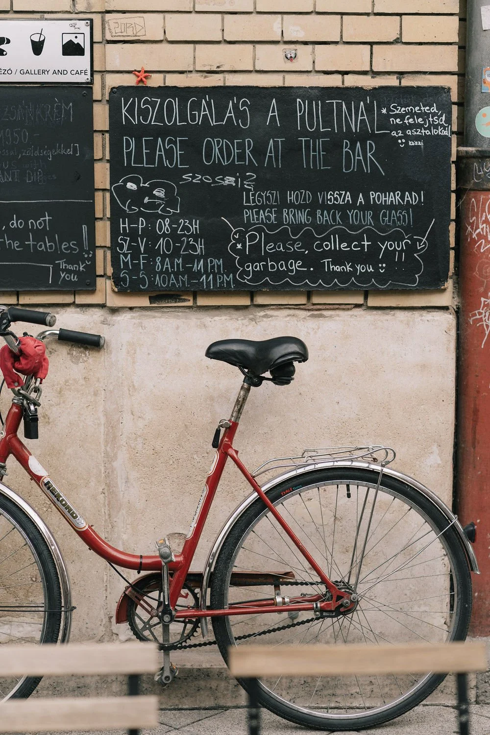 Quadro de avis em um muro de tijolos, solicitando pedidos no bar e informações de horário, com uma bicicleta vermelha estacionada na rua.