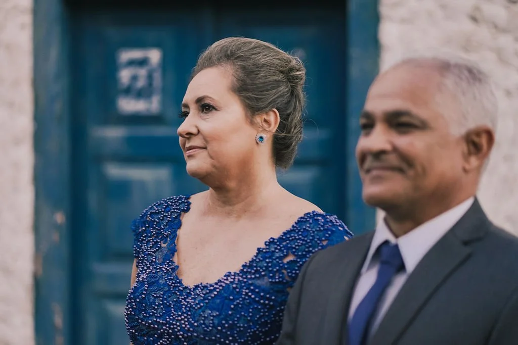 Mulher idosa com cabelo cinza em um vestido azul com detalhes perolados, ao lado de um homem idoso com cabelo branco em terno e gravata, ambos sorrindo, na frente de uma parede de pedra e uma porta azul ao fundo.