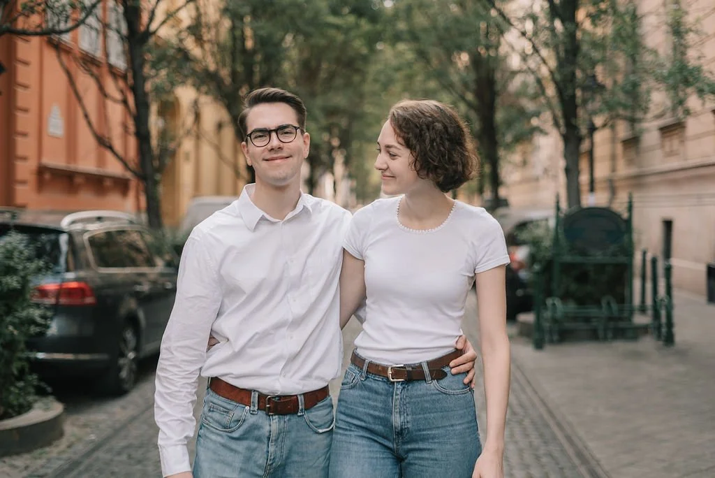 Um casal de jovens caminhando na rua, sorrindo, com árvores e carros ao fundo, em um ambiente urbano
