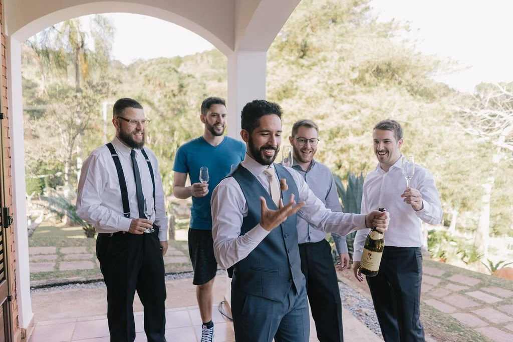 Homens vestindo roupas formais, celebrando com taças de champanhe em uma varanda ao ar livre, com um homem no centro segurando uma garrafa de champagne.