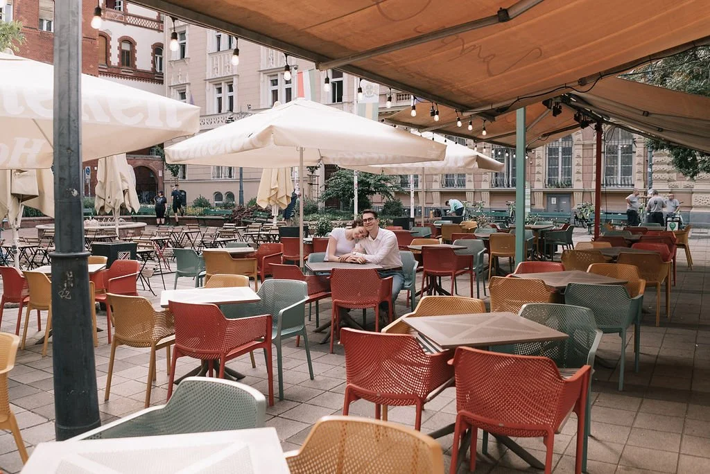 Pessoa sentada em uma mesa ao ar livre em um café com cadeiras coloridas, guarda-sóis e iluminação pendurada, em uma praça urbana.