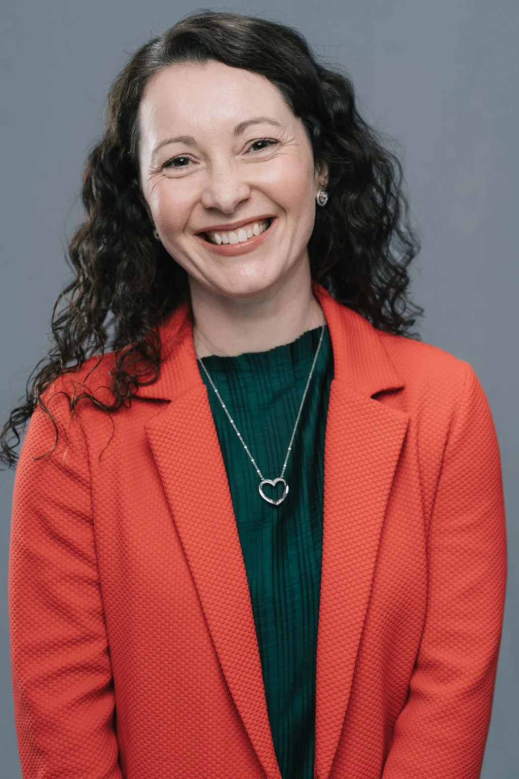 Mulher sorrindo, com cabelo cacheado, usando um blazer laranja e uma camiseta verde, com um colar de coração prateado, contra um fundo cinza.
