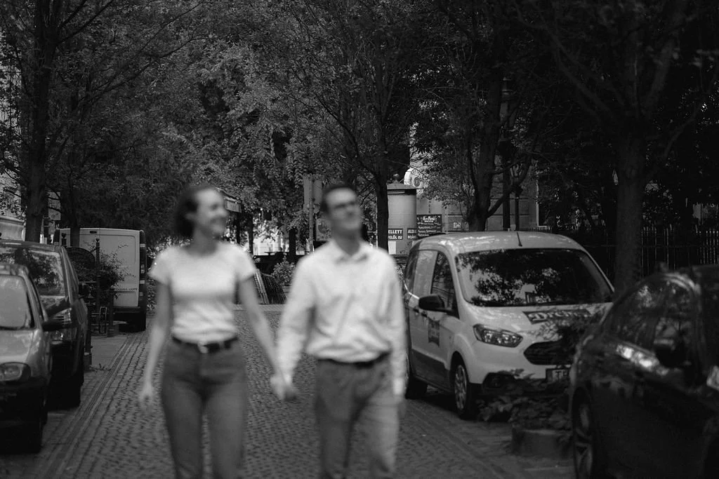 Casal caminhando de mãos dadas na rua, ao entardecer, em uma cidade. Estacionamento com vários carros e árvores ao fundo.