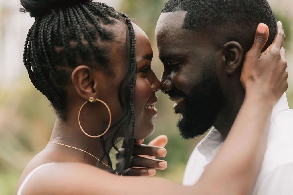 Um casal se encarando com sorrisos e concentração, segurando um ao outro, em um ambiente ao ar livre.