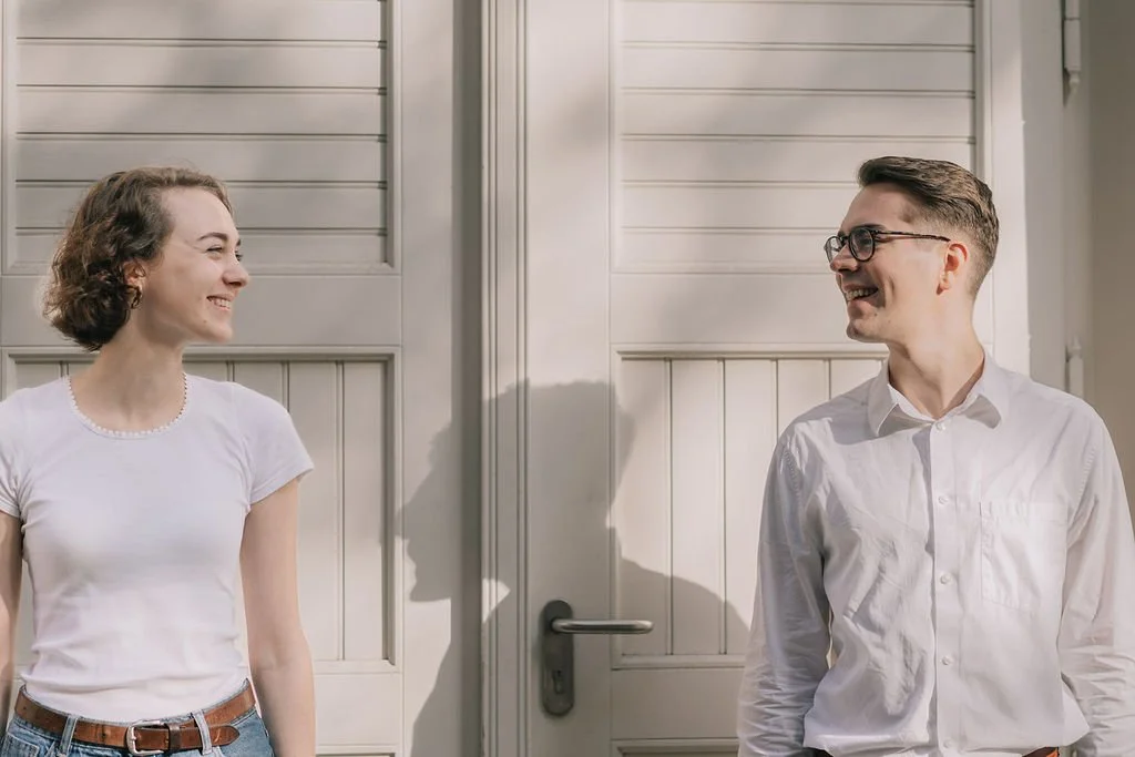 Um homem e uma mulher sorrindo um para o outro em frente a uma porta branca, com luz natural. Ambos usam roupas brancas e parecem estar felizes.