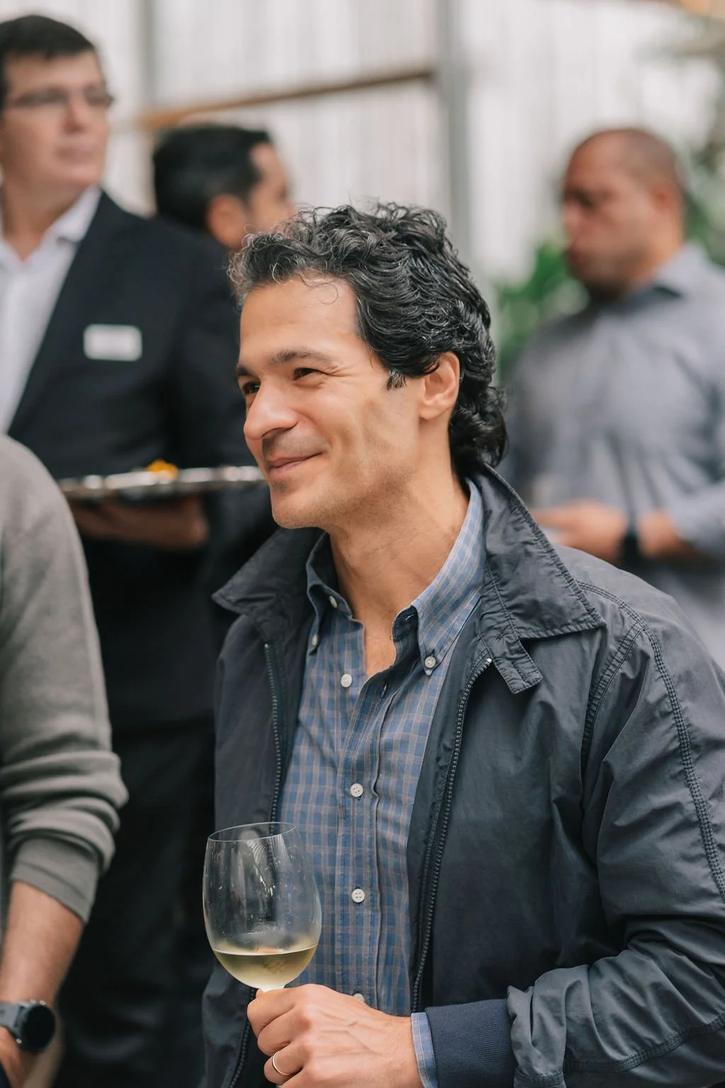 Homem sorrindo com taça de vinho branco em um evento social, ao fundo outras pessoas conversando.