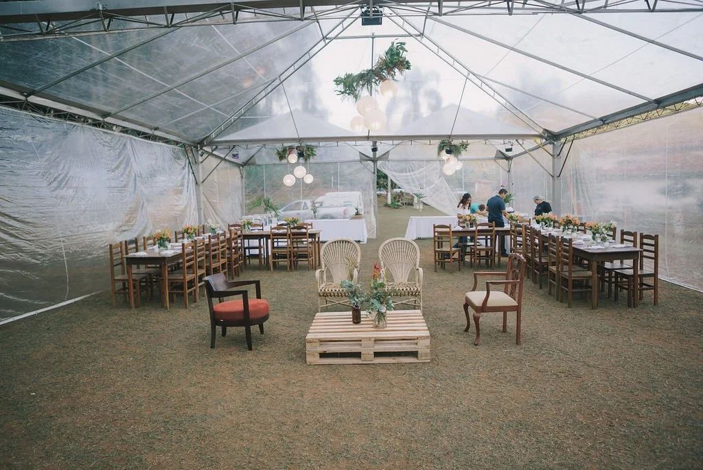 Decoração de uma festa ao ar livre dentro de uma tenda transparente, com mesas e cadeiras de madeira, iluminação pendurada e arranjos de flores, pronto para um evento.