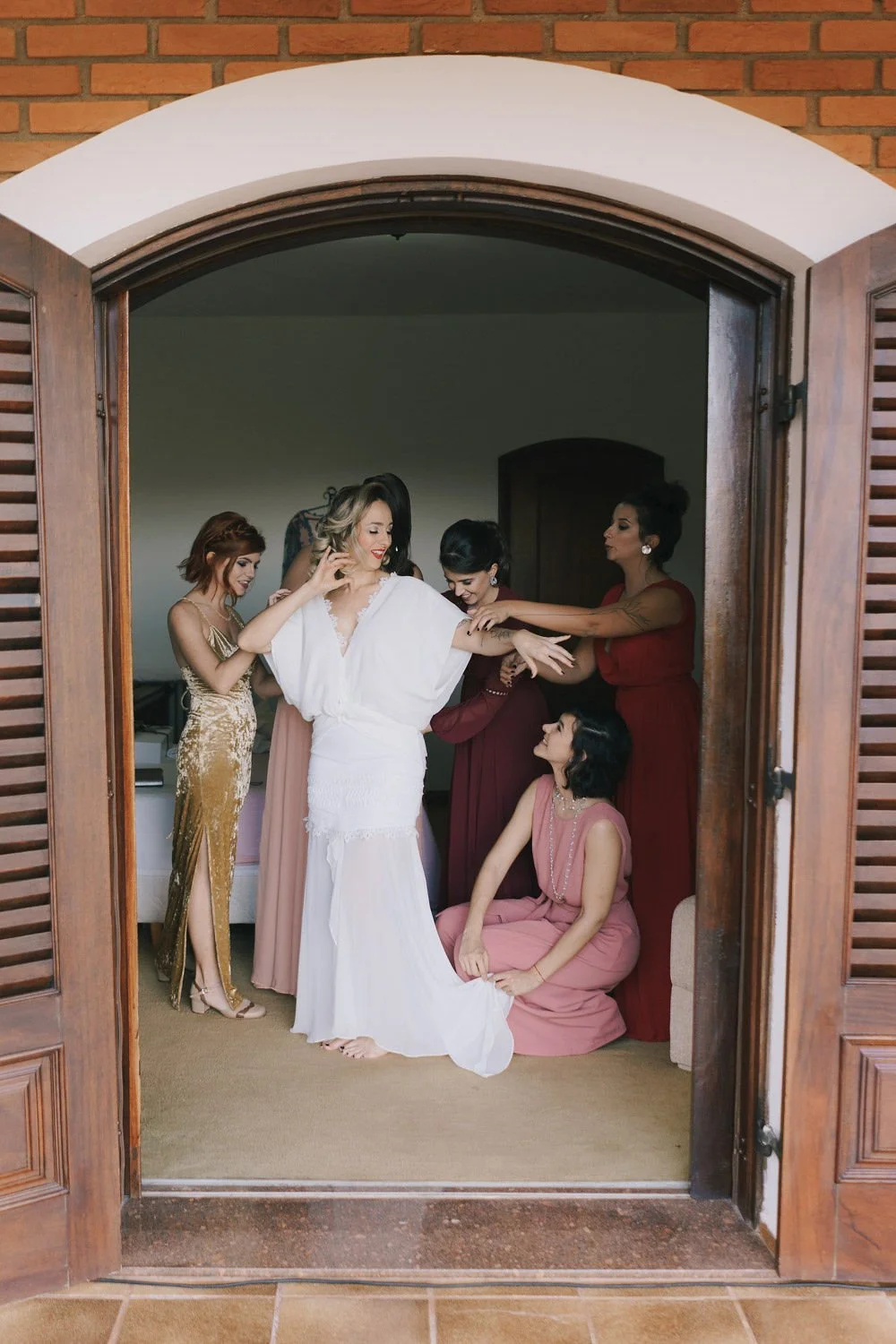 Grupo de mulheres ajudando a noiva a se vestir em um quarto, vistas pelo vão de uma porta de madeira