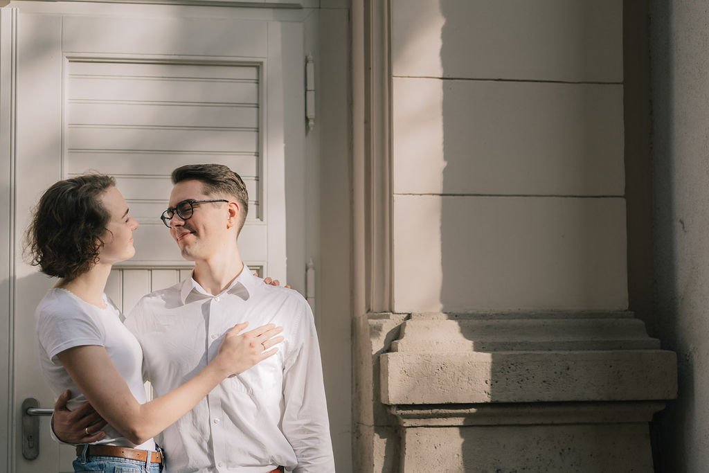 Um casal sorrindo um para o outro, de frente, em uma porta de entrada de uma casa ou prédio, com luz natural e sombra na parede ao lado.
