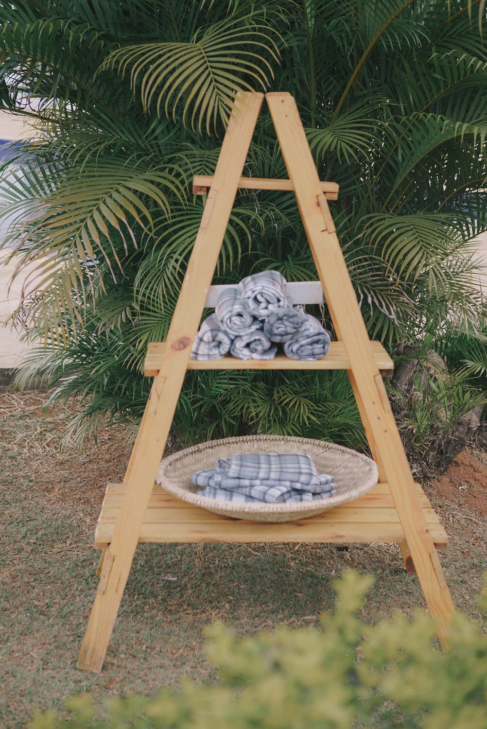 Estante de madeira em formato de escada com roupa de cama de padrão de risca cinza e branca, colocado ao ar livre com plantas verdes ao fundo.