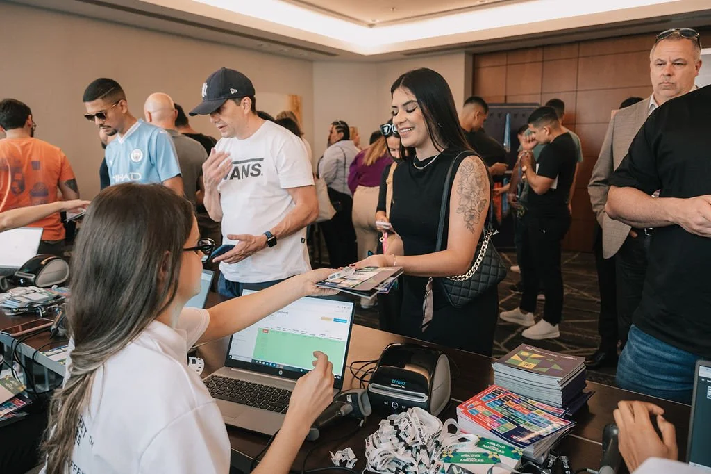 Mulher sorridente recebendo um flyer de uma atendente em um evento, com várias pessoas ao fundo.