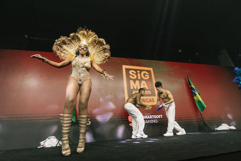 Uma mulher vestida com uma fantasia dourada com penas e uma saia de salto alto, posando com braços abertos em um palco. Ao fundo, duas pessoas dançando, uma segurando um prêmio, com fundo de uma tela que exibe 'SIGMA AWARDS ÁFRICAS' e a bandeira do B