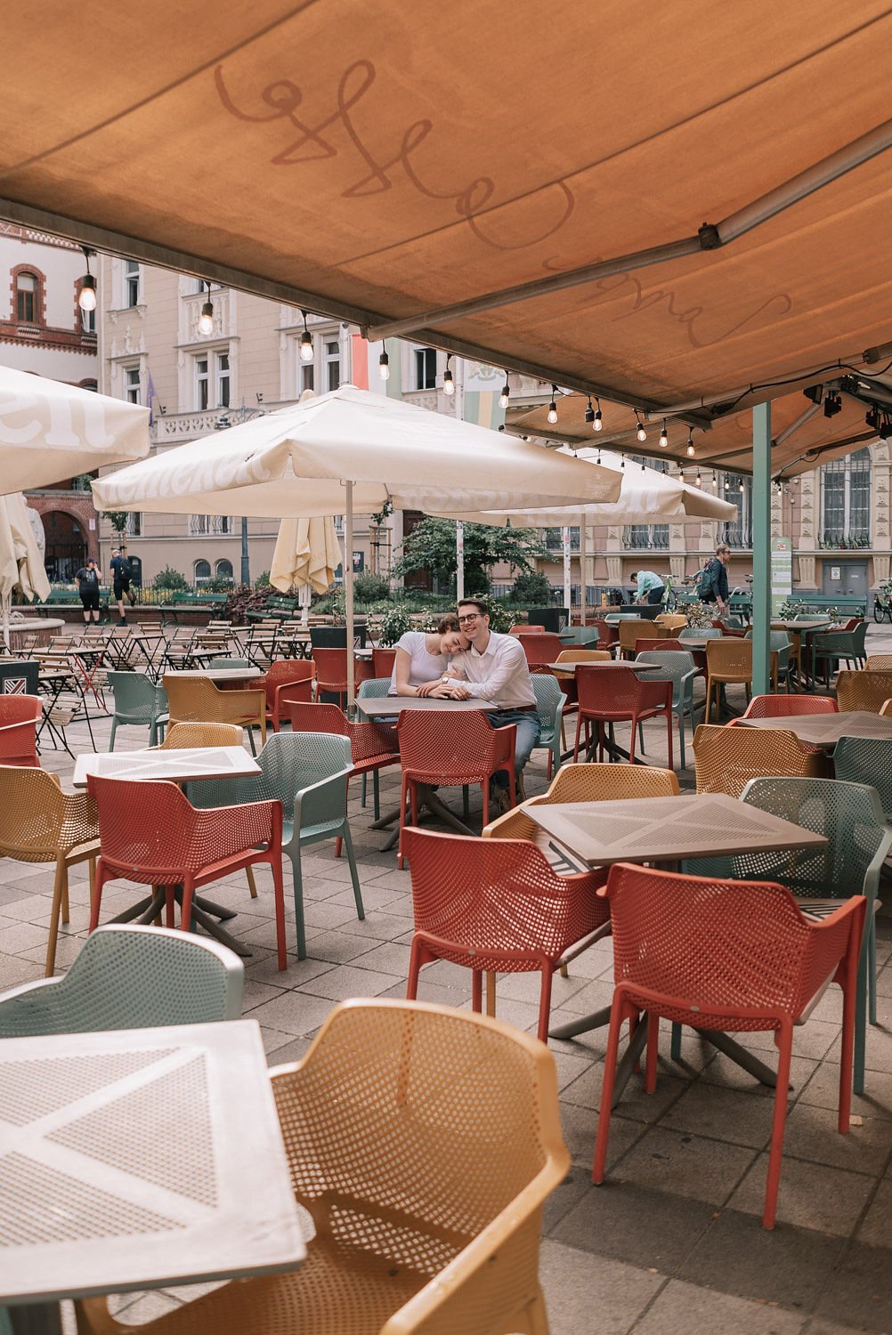Casal sentado em uma mesa ao ar livre de um café ou restaurante, cercado por cadeiras coloridas e ombrelones.