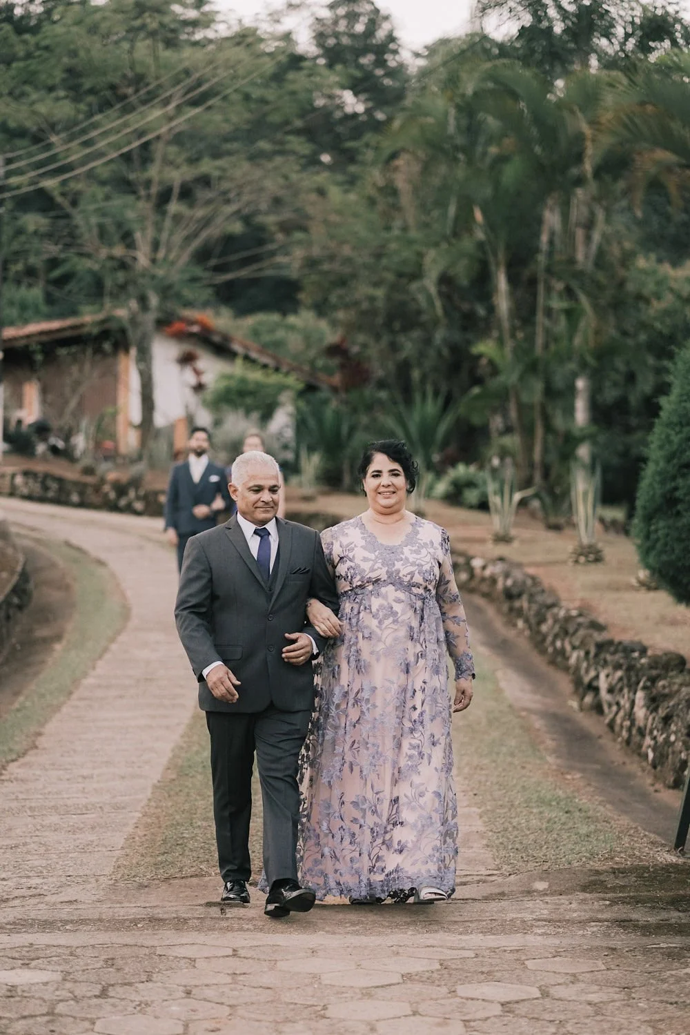 Casal caminhando ao ar livre, com árvores e casa ao fundo, rua de pedra.
