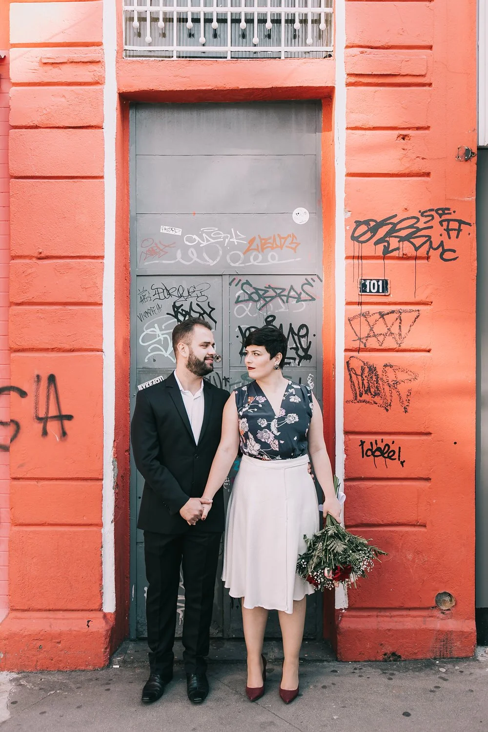 Casal segurando mãos na rua, com mulher segurando buquê de flores, em frente a parede laranja com graffiti, R$ 101 na placa
