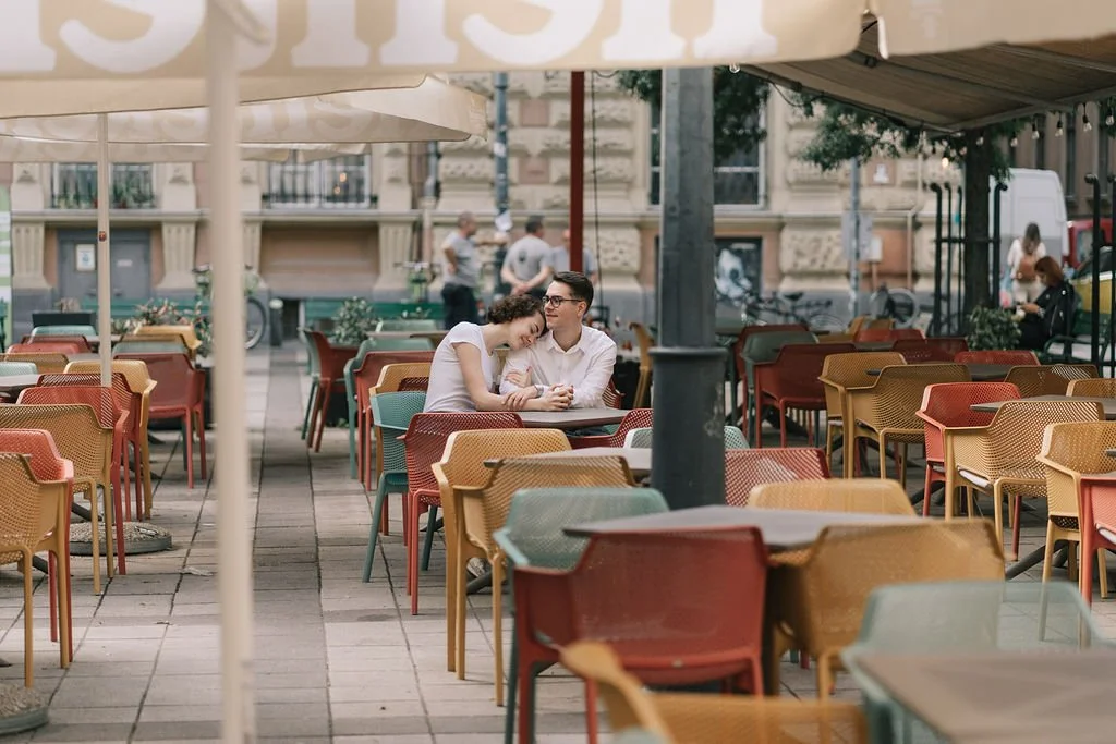 Casal sentado em uma mesa ao ar livre de um café ou restaurante, sob guarda-sóis em uma área urbana.