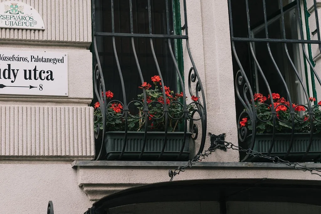 Janela com grades decorativas de ferro e vasos de flores vermelhas na varanda de um prédio com placas de rua e sinalização em húngaro e português.