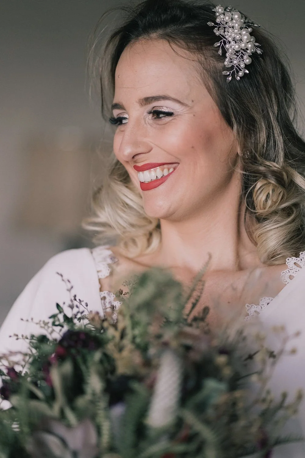 Mulher sorrindo de perfil, com cabelo cacheado loiro, usando uma tiara de pérolas e maquiagem elegante, segurando um buquê de flores.