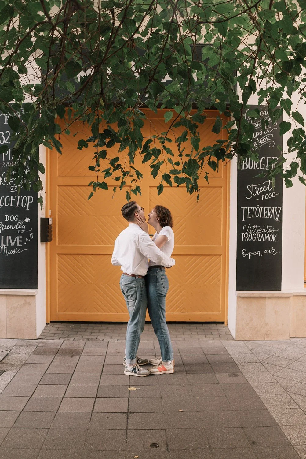 Casal se abraçando e beijando na frente de uma porta laranja, com um galho de árvore caindo sobre eles. Ao redor, quadros de lousa com desenhos e frases em inglês sobre comida, música e eventos ao ar livre.