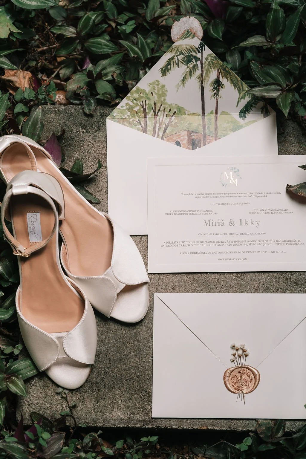 Convite de casamento, sapatos de noiva, cartão convite e envelope com lacre de cera, tudo disposto sobre uma pedra com folhas verdes ao redor.