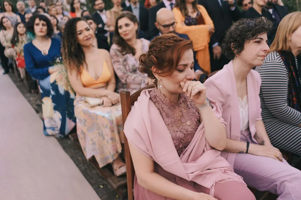Mulher chorando em uma cerimônia com pessoas ao redor. Ela está com as mãos na boca, demonstrando emoção.