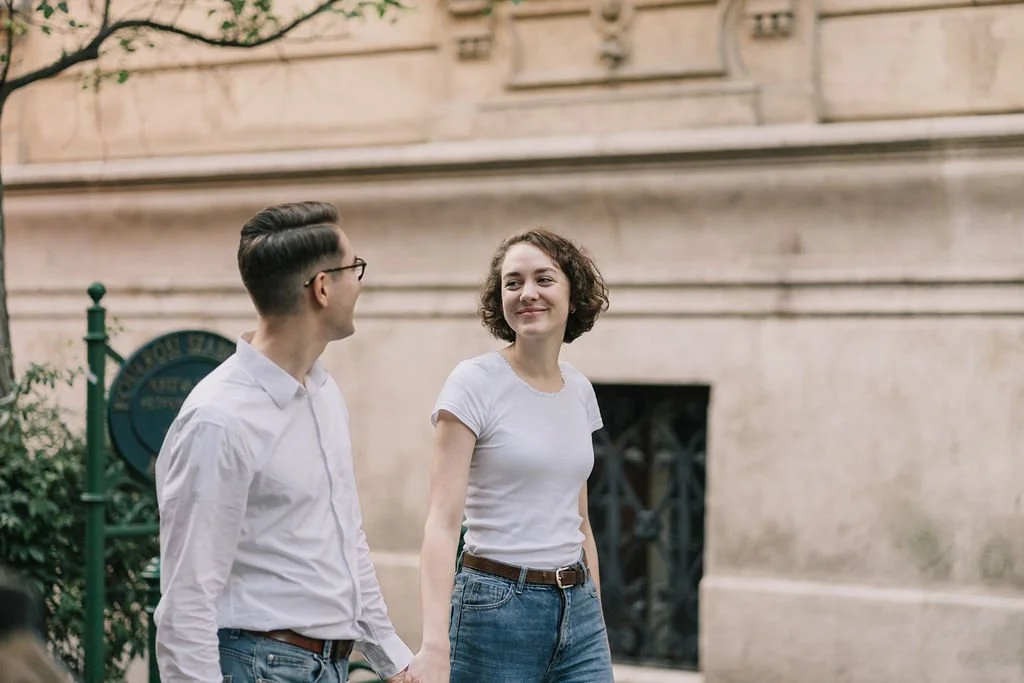 Casal sorridente caminhando na rua, com árvores e parede de edifício ao fundo.