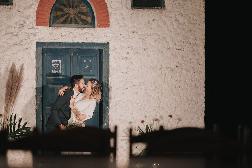 Casal apaixonado dançando na frente de uma porta azul com parede de textura rústica, durante a noite.
