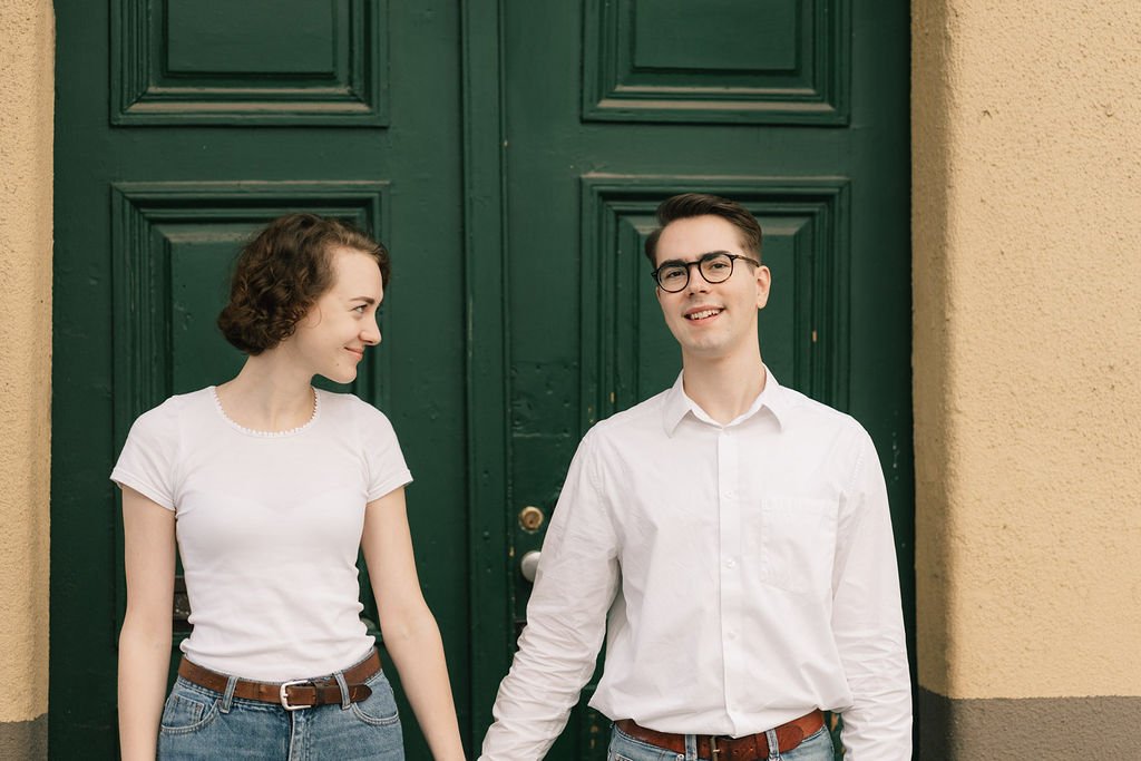 Um jovem casal segurando as mãos em frente a uma porta verde, sorrindo um para o outro.