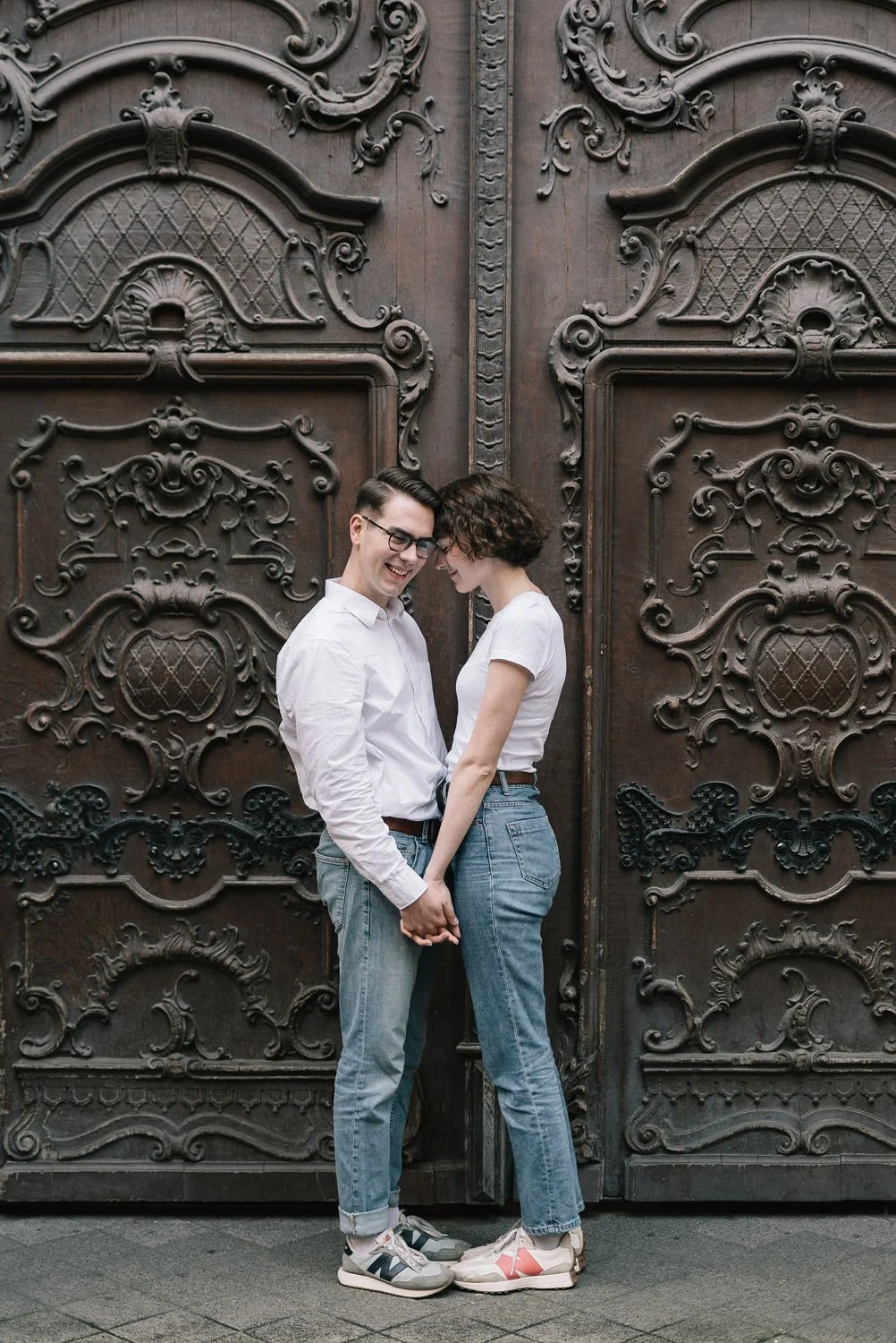 Casal sorridente segurando as mãos diante de uma grande porta de madeira com detalhes ornamentais.