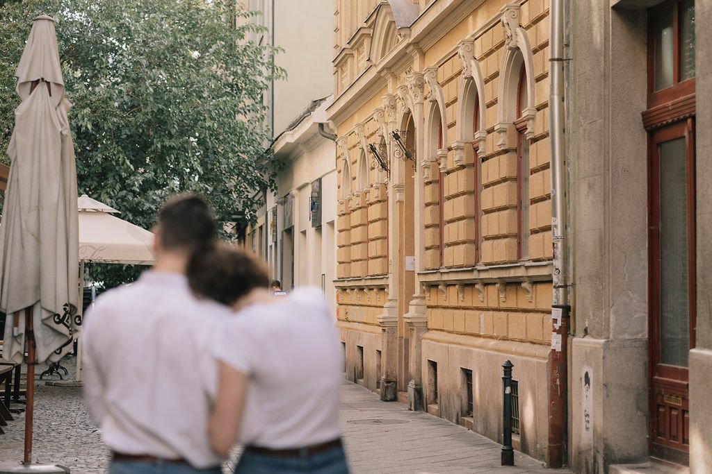 Rua com prédios históricos de arquitetura clássica, detalhes ornamentais, janelas arqueadas e paredes em tons de bege. Pessoas de costas conversando na calçada, árvores e guarda-sóis ao fundo.