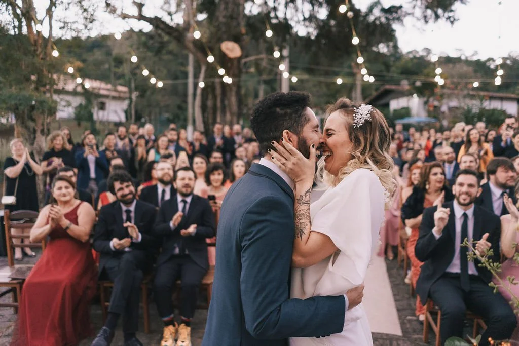 Casal de noivos se beijando no altar ao ar livre durante cerimônia de casamento, com convidados aplaudindo ao fundo.