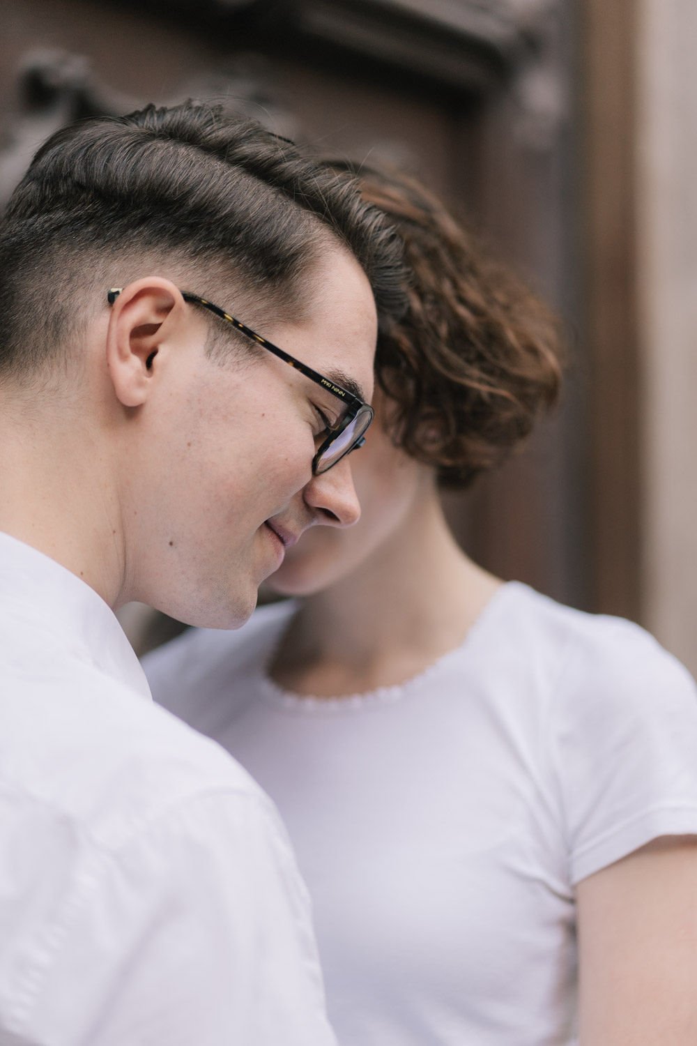 Close-up de duas pessoas sorrindo, uma usando óculos e com cabelo curto e penteado com laterais raspadas, a outra com cabelo castanho ondulado e cabelo curto com cabelos cacheados, ambas vestindo camisetas brancas, em um ambiente interno.