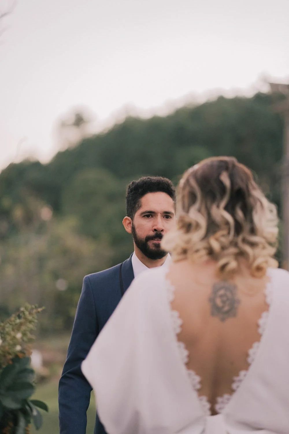 Homem de terno azul observando uma mulher com cabelo cacheado loiro, usando vestido branco com detalhes de renda, ao ar livre com árvores ao fundo.
