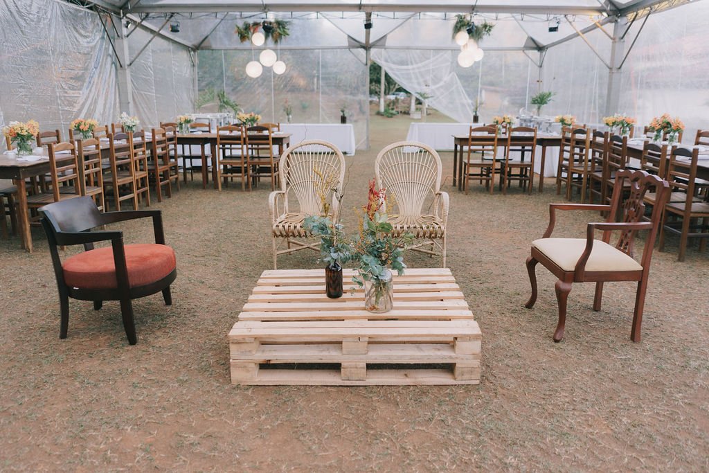 Decoração de ambiente de casamento ou evento ao ar livre com cadeiras, mesas, plantas e uma estrutura de tenda transparente.