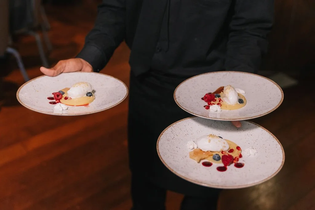 Garçom segurando três pratos de sobremesa decoradas com sorvete, frutas vermelhas, blueberries e creme, em ambiente de restaurante.