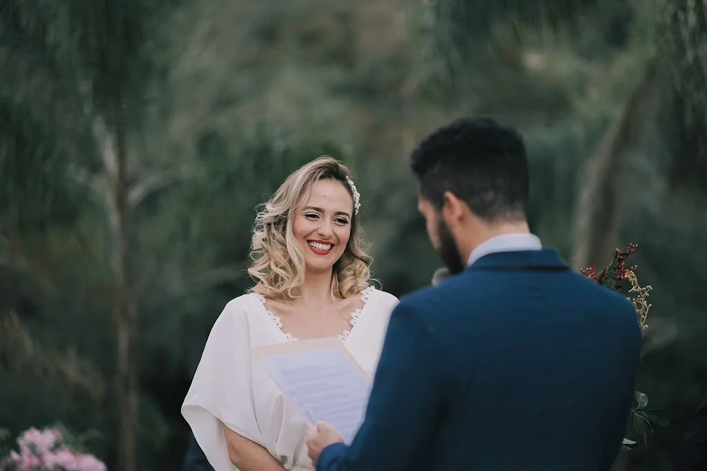 Casal no casamento ao ar livre, o homem de costas segurando uma folha de papel, a mulher sorrindo Feliz.