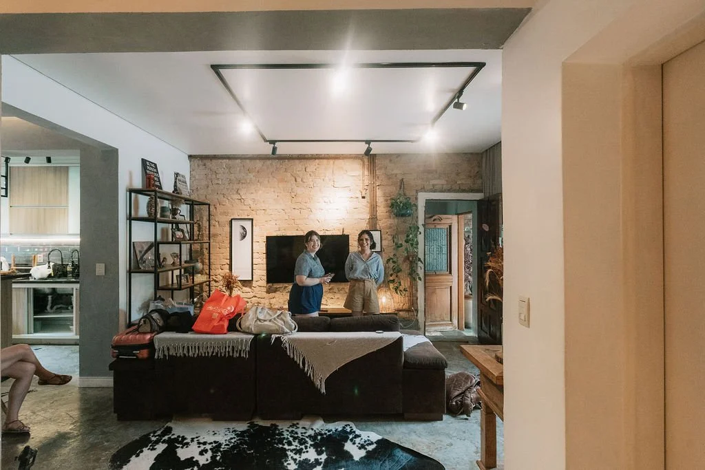 Sala de estar com dois homens de pé na frente de uma parede de tijolos expostos e sofá marrom, com uma estante de livros ao lado esquerdo, uma mulher na cozinha à esquerda, e uma porta de madeira ao fundo à direita.