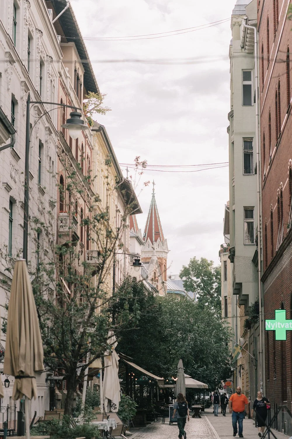 Rua com prédios de arquitetura antiga, árvores e pessoas caminhando, com igreja de torre pontiaguda ao fundo.