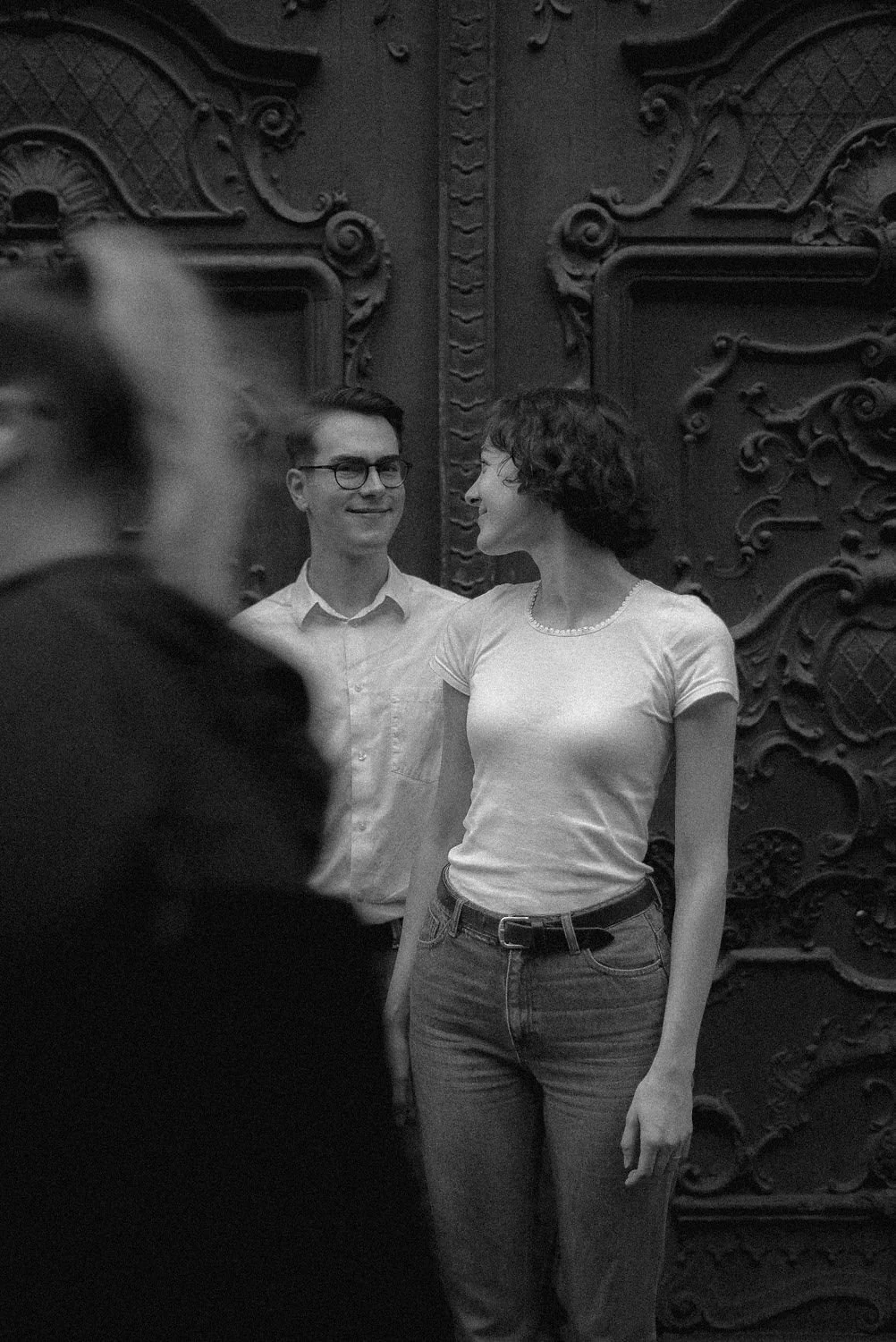 Homem e mulher sorrindo um para o outro em frente a uma porta de madeira com detalhes decorativos, em um ambiente interno.