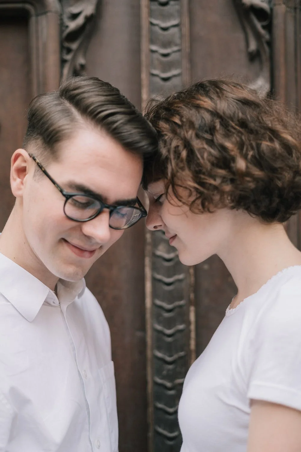 Um casal emparelhado com testa encostada, olhos fechados, sorrindo suavemente, em um momento íntimo, com fundo de madeira decorativa.