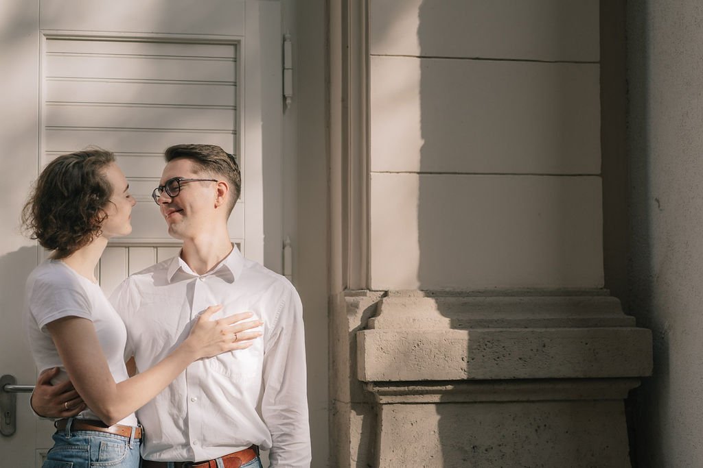Um casal de jovens sorrindo e olhando um para o outro na rua, ao lado de uma parede de prédio com sombra.
