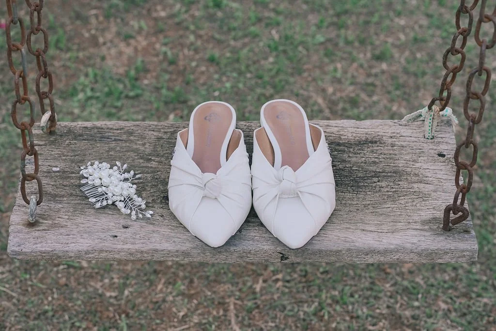 Par de sapatos brancos de salto baixo com laços, uma pinha com pérolas e detalhes de renda, colocado em um balanço de madeira.