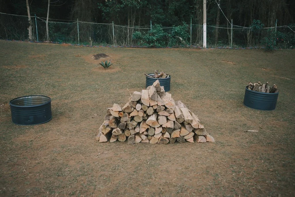 Pilhas de lenha empilhadas ao centro de um espaço ao ar livre, cercado por cercas, com baldes contendo mais lenha ao redor.