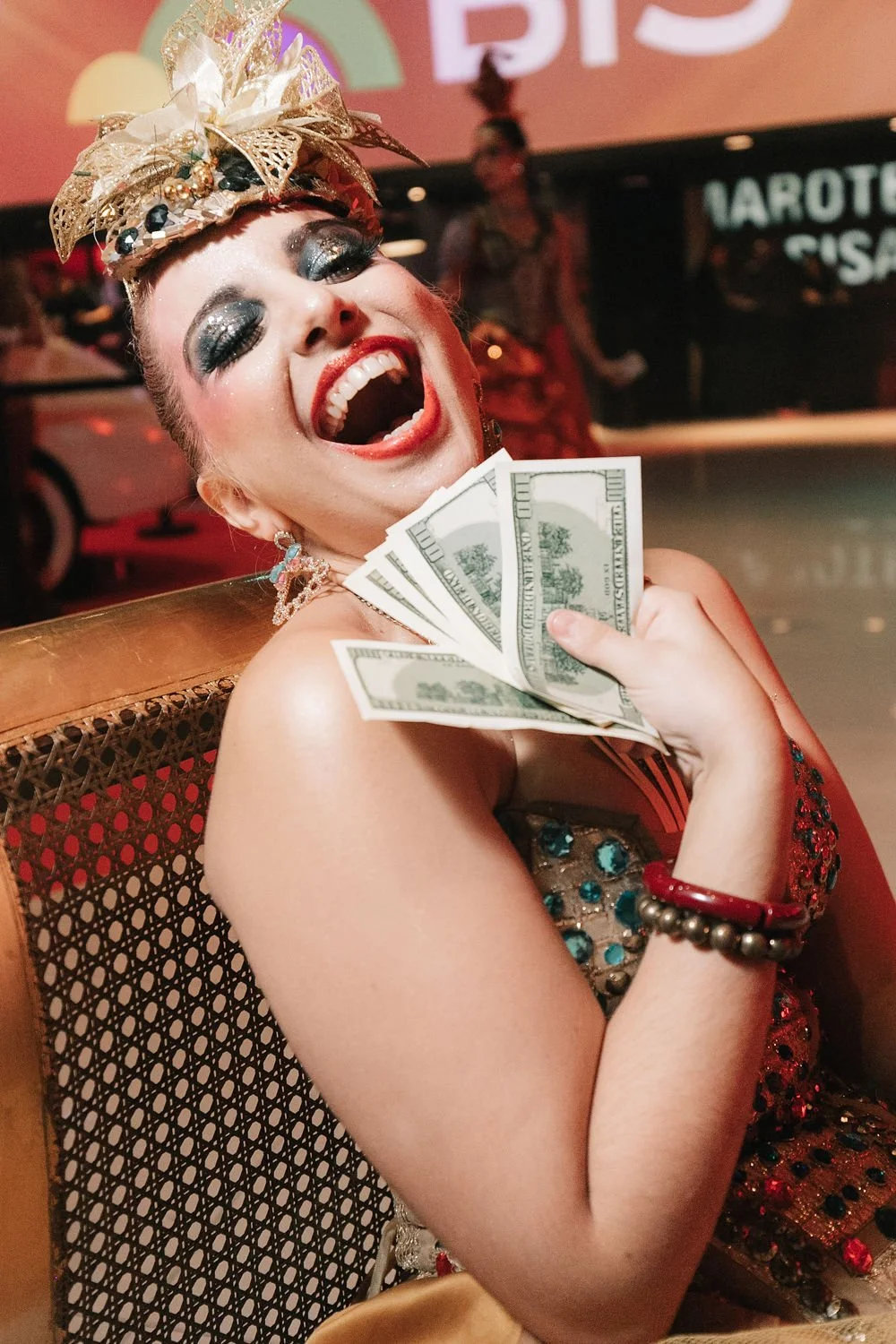 Mulher sorrindo e celebrando com dinheiro em mãos em um ambiente de festa, usando maquiagem marcante e acessório de cabeça ornamentado.