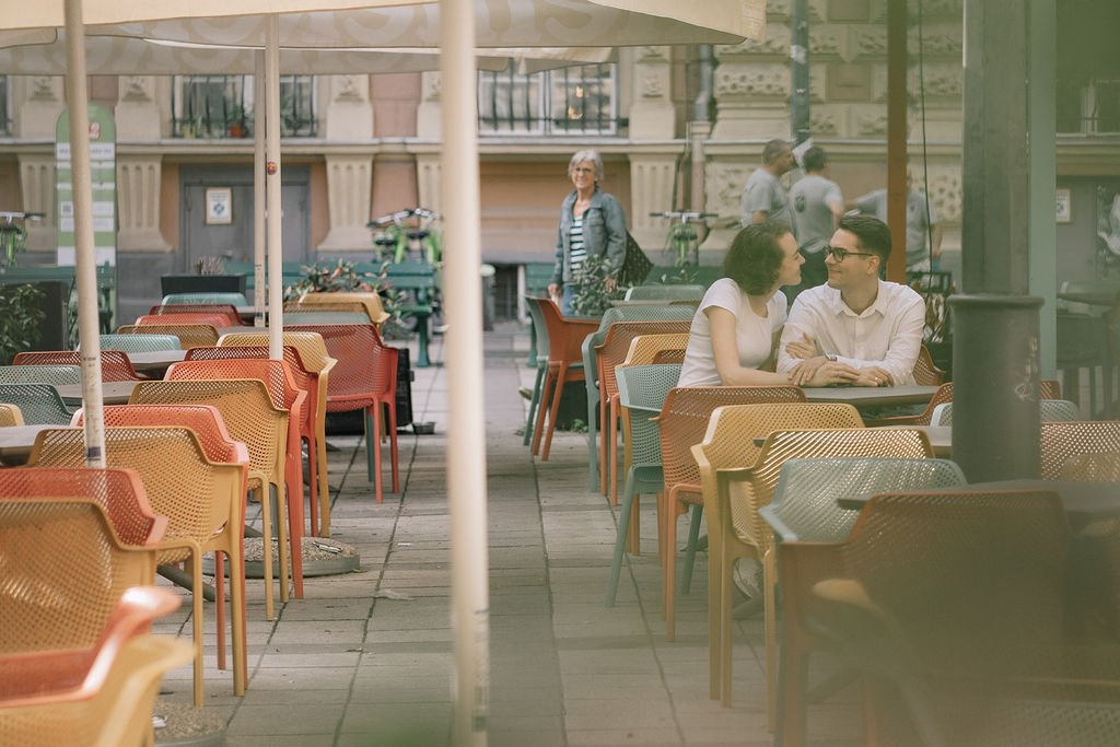 Homem e mulher conversando sentados em uma mesa ao ar livre de um restaurante, com outras mesas vazias ao redor e uma pessoa mais atrás sorrindo.