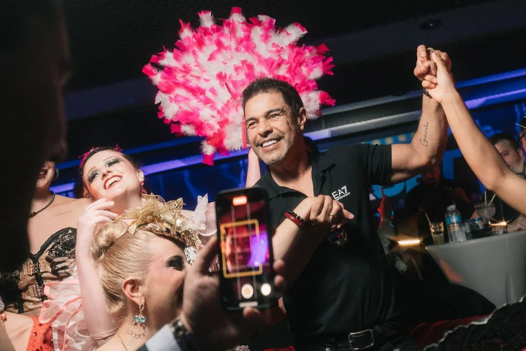 Homem sorridente dançando com mulheres vestidas com fantasias e um vestido de baile, enquanto outra pessoa grava a cena com o celular, em um ambiente de celebração com iluminação colorida.
