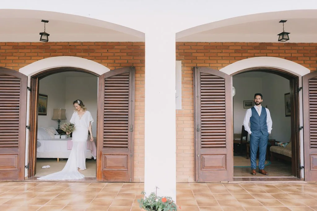 No lado esquerdo, uma noiva de vestido branco segurando um buquê, dentro de um ambiente com parede branca, uma luminária e uma cama. No lado direito, um homem de terno azul escuro e camisa branca dentro de uma sala com paredes brancas, poltrona e mes