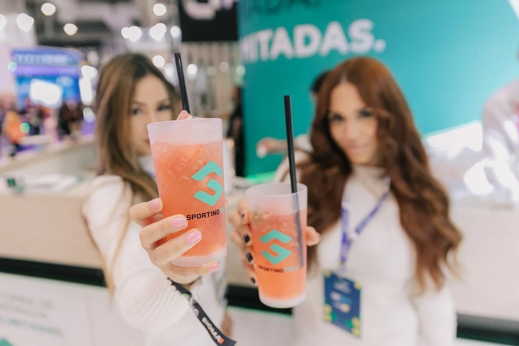 Duas mulheres segurando copos de bebida rosa com canudos, em um evento com sinalização de fundo, ambas vestindo roupas claras.