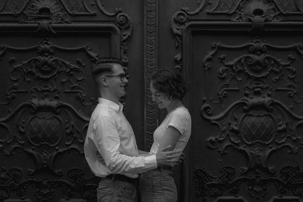 Um casal jovem sorrindo e segurando um ao outro na frente de uma porta de madeira decorada.